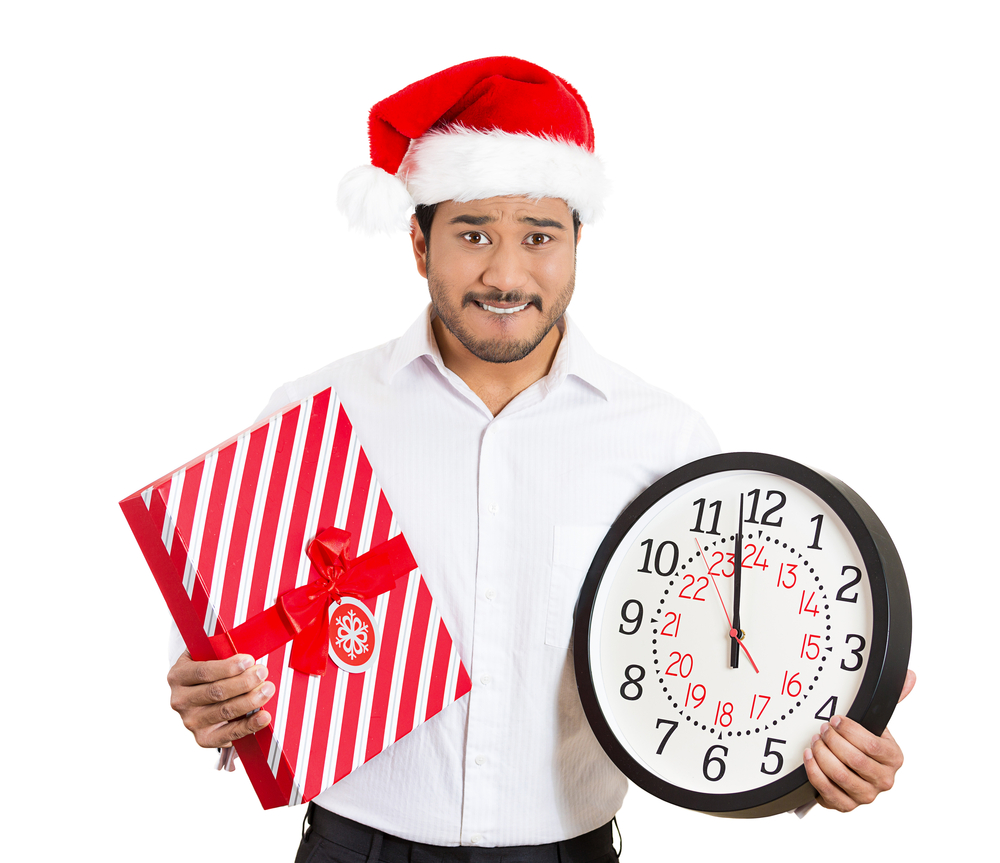 Closeup portrait of worried young man wearing red santa claus hat, holding clock and gift in hands, isolated on white background. Negative emotion facial expression. Last minute christmas shopping